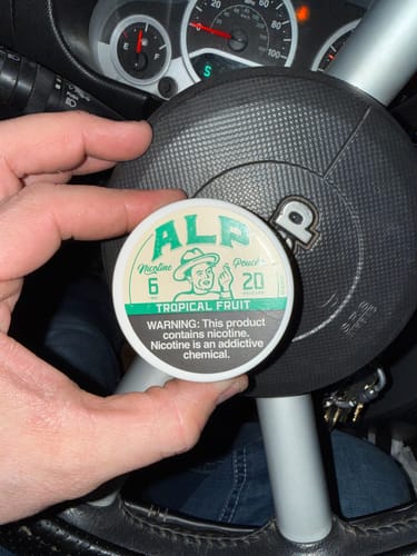 Customer holding a can of ALP Tropical Fruit Nicotine Pouches in front of a car's steering wheel.