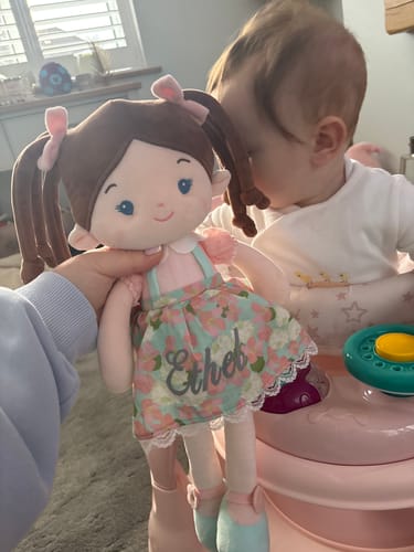 Customer holding a personalized Leya Doll with 'Ethel' on its floral dress, as a baby looks on in the background.