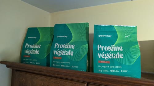 Un client montre ses trois sachets verts de Protéine Végétale Bio sur un meuble en bois.