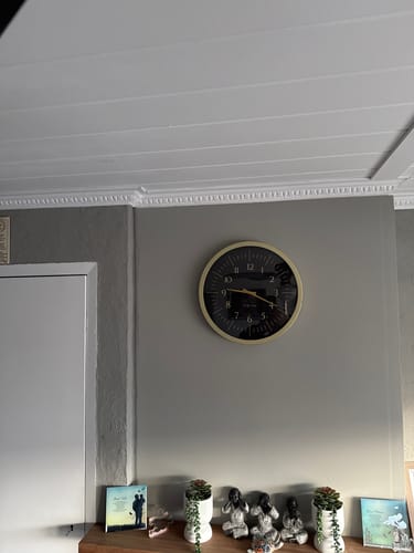 Klant toont de goud-zwarte NeXtime 'Galaxy' wandklok die aan een grijze muur hangt.