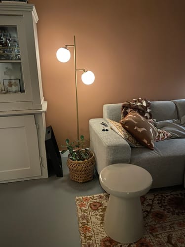 Klant toont de verlichte gouden Pallon vloerlamp met twee bollen naast een bank in een woonkamer.