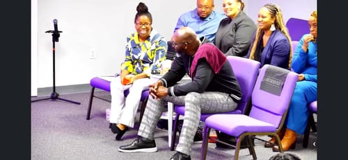 Customer and her husband seated at an event, smiling in anticipation of their custom anniversary song.