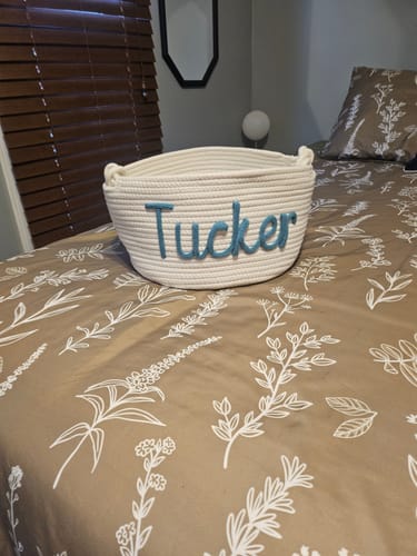 Customer's white Personalized Knitted Basket with the name 'Tucker' in blue yarn, sitting on a bed.
