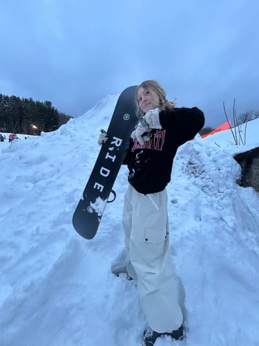 Customer standing in the snow with a snowboard, wearing the beige baggy waterproof snow pants.