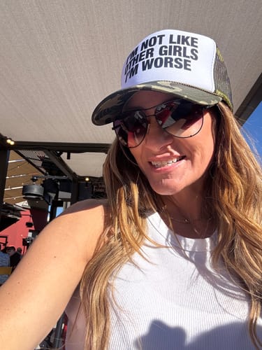 Reviewer smiling while wearing the 'I'm Not Like Other Girls I'm Worse' trucker hat with a camo brim outdoors.