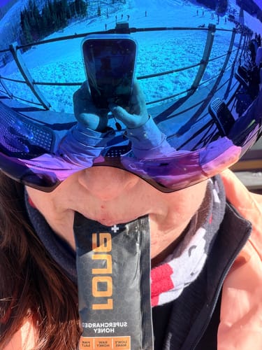 Close-up of a reviewer in ski goggles eating a SuperCharged Honey packet on a snowy mountain.