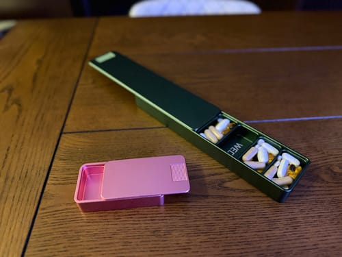 Customer's two metal pill cases, a small pink one and a long green one, open on a wooden table.