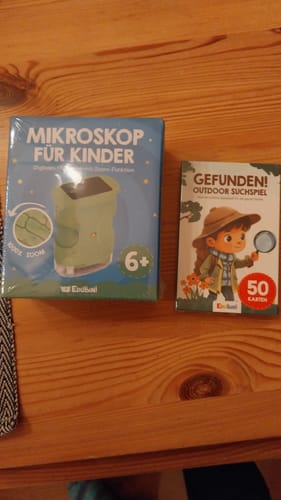 Rezensent zeigt die Verpackung des Edubini Gefunden! Suchspiels neben einem Kindermikroskop auf einem Holztisch.