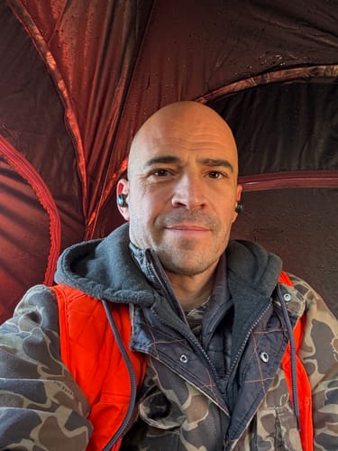 Reviewer wearing the black XCOR PRO earbuds while sitting inside a hunting blind in camouflage gear.