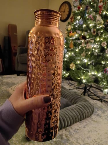 Customer's hand holding the shiny, hand-hammered Pure Copper Water Bottle in a living room.