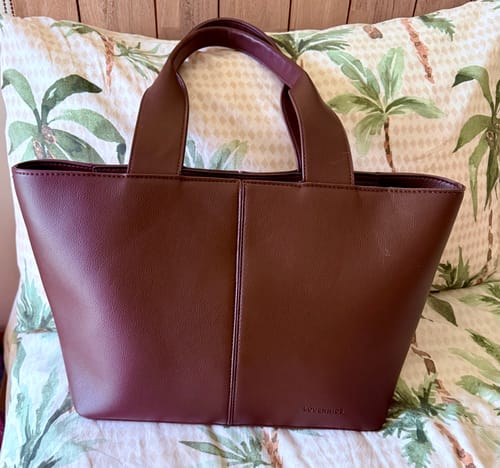 Reviewer's chocolate Henley Tote Bag resting against a pillow with a palm tree pattern.