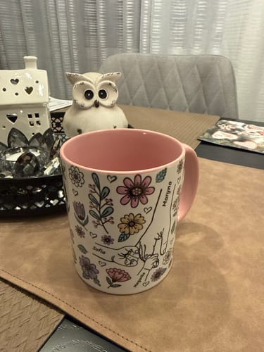 Kundens Kärleksmugg med rosa insida och blommigt tryck med händer och namn, står på ett bord.