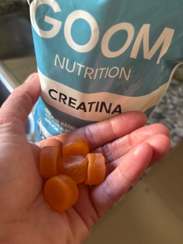 Cliente sosteniendo varias gomitas de creatina naranjas en la mano, con el empaque del producto al fondo.