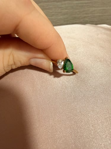 A customer's hand holds the Leesha Birthstone Ring, featuring a green pear-shaped stone next to a clear heart-shaped stone.