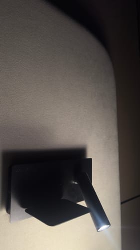 Customer's black Kross wall reading lamp mounted on a headboard and switched on in a dim room.