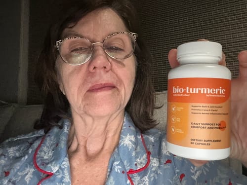 Reviewer in pajamas holding up a white and orange bottle of Bio Turmeric Elite supplements.