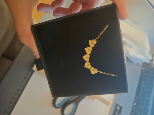 Reviewer holds a black box, showing the gold Little Heart necklace with five initial pendants.