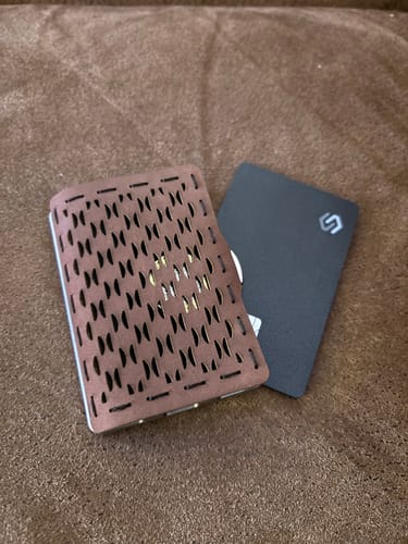 Customer's black SPOTMINDERS Tracking Card PRO shown next to a brown, perforated wallet on a soft surface.