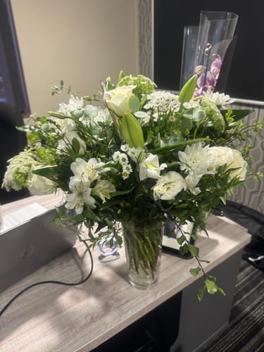 Reviewer's White Purity Vase Arrangement with full white flowers and greenery in a clear vase on a desk.