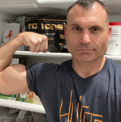 Reviewer flexing his bicep with a pouch of SuperCharged Honey visible on a shelf in the background.