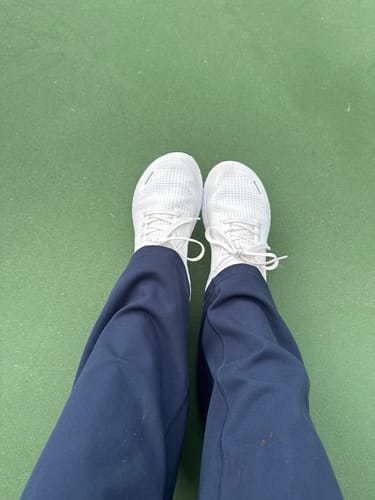 Reviewer's point of view looking down at their white MUUV Stride Pickleball Shoes on a green court.