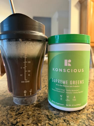 Customer's tub of Supreme Greens next to a shaker cup filled with the prepared, frothy green drink on a kitchen counter.