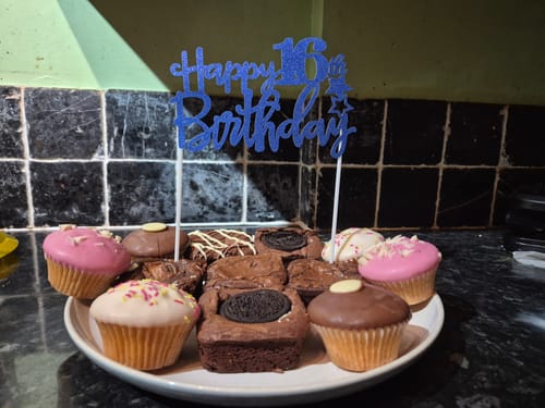 Customer's mixed box of postal brownies on a plate with cupcakes and a blue 'Happy 16th Birthday' topper for a celebration.