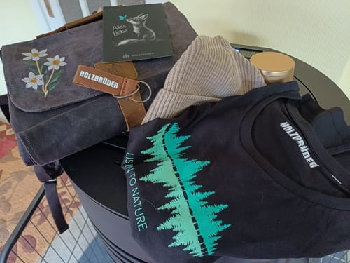 Kunde zeigt den Inhalt seiner Holzbrüder® Wundertüte: eine graue Tasche, ein schwarzes T-Shirt, eine Mütze und Zubehör.