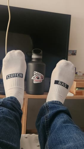 Customer relaxing with their feet up, wearing white TUNERCULT CREW SOCKS with 'CLUTCH' and 'GAS' text on them.
