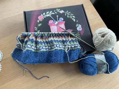 Reviewer's in-progress fair isle knitting project from the Ladies Mystery Knitting Box, with yarn and the box on a table.