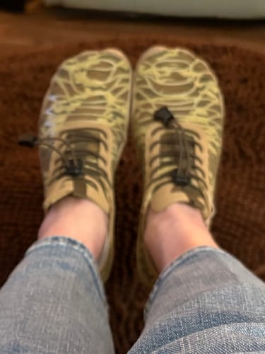 Close-up: Reviewer wearing khaki Lorax Pro barefoot shoes with a yellow abstract pattern on a brown rug.