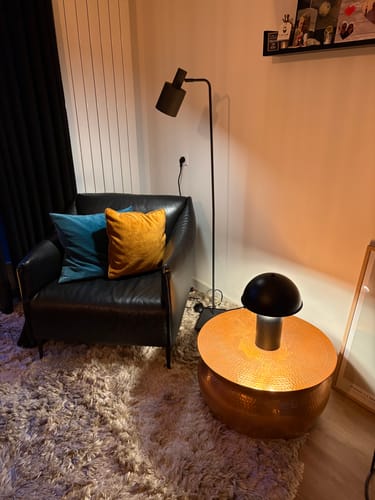 Klantfoto van de verlichte, zwarte Bulb Mushroom tafellamp op een koperen bijzettafel in een woonkamerhoek.