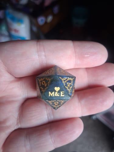 Customer's hand holding a personalized D20 die, showing the initials 'M & E' and a heart in gold.