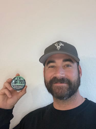 A smiling reviewer holding up a round tin of Refreshing Chill Nicotine Pouches against a white wall.