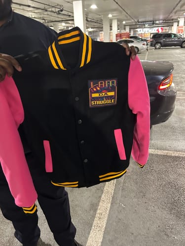 Customer holds a custom varsity jacket with a black body, pink sleeves, and a colorful chest patch in a parking garage.