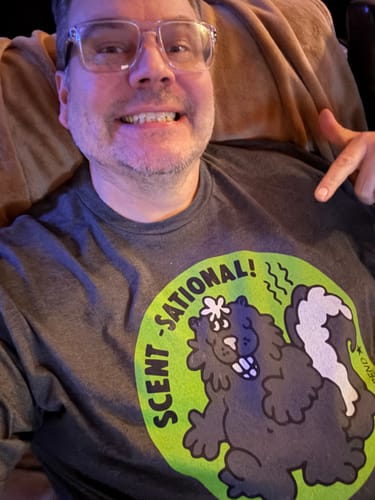 Reviewer smiling while pointing to the skunk graphic on their grey Scent-Sational Scratch N Sniff T-Shirt.