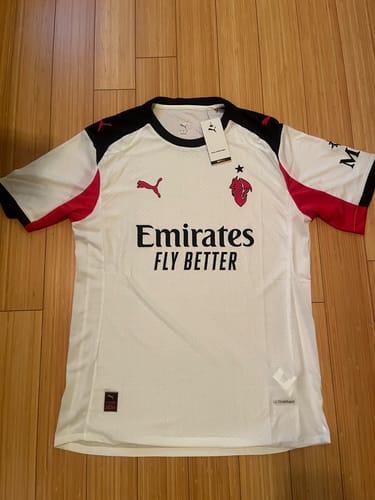Un recensore mostra la maglia da trasferta del Milan 2025/26 stesa su un pavimento di legno.