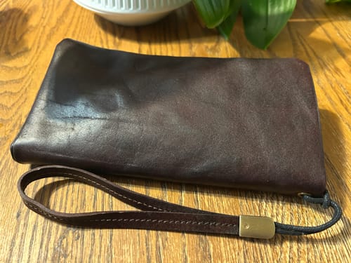 Customer's soft brown leather wallet with attached wrist strap, shown lying flat on a wooden table.