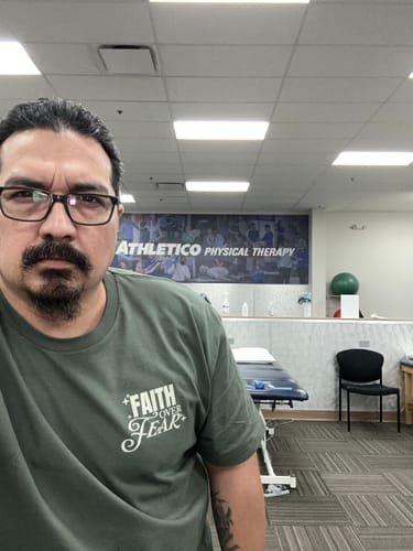 Reviewer wearing the green COURAGE TEE with a 'Faith over Fear' graphic in a physical therapy office.