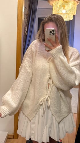 Kundin macht ein Spiegelselfie und trägt den cremefarbenen Oversized Cardigan mit Schleifen über einem weißen Kleid.