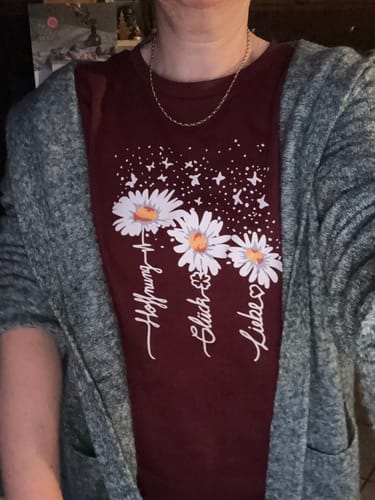 Kundin trägt das burgunderrote 'Hoffnung, Glück, Liebe' Shirt mit Gänseblümchen-Motiv unter einer grauen Strickjacke.