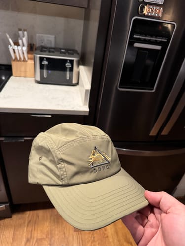Customer's hand holding the light green Big Camp Hat Ripstop in a kitchen, showing the front logo and 5-panel design.