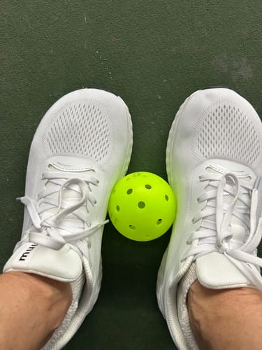 Reviewer's feet in white MUUV Flow pickleball shoes, with a yellow pickleball between them on a court.