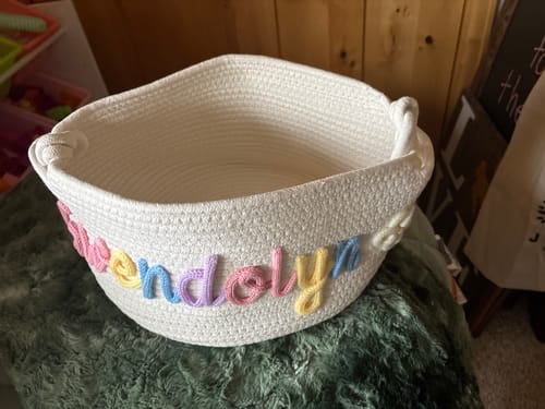 Customer's personalized off-white knitted basket with the name 'Gwendolyn' in colorful pastel yarn on a green surface.