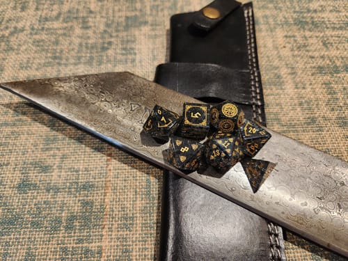 Customer's Nordic Black Shattered Glass Dice Set with gold details displayed on the blade of a wide, patterned knife.