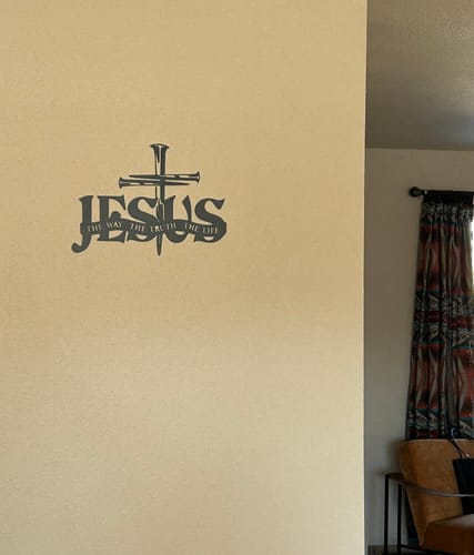 Customer's black Jesus & Cross metal wall decor hanging on a beige wall in their room.