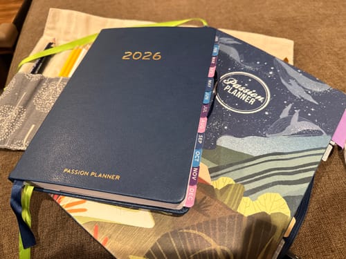 Customer's navy blue 2026 Weekly Hardcover Planner resting on another planner, with colorful monthly tabs visible on the side.