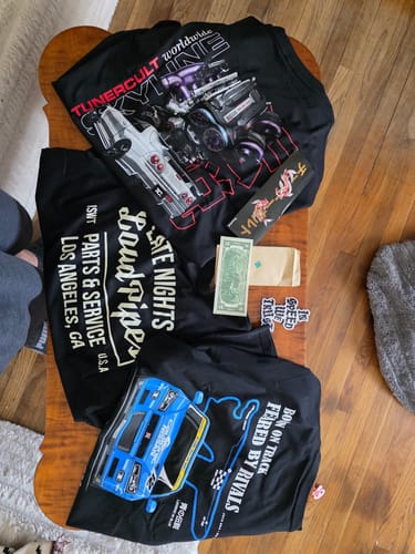 Customer shows the contents of their TCG90 MYSTERY CASH BOX: three black graphic t-shirts, a sticker, and a two-dollar bill.