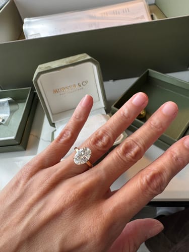 Customer's hand wearing the large oval solitaire diamond ring with a gold band, with the open jewelry box in the background.