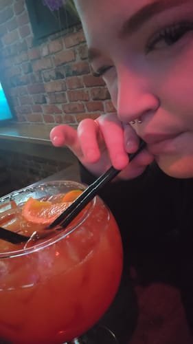 Close-up of a reviewer sipping a large orange cocktail from a fishbowl glass in a dimly lit bar.